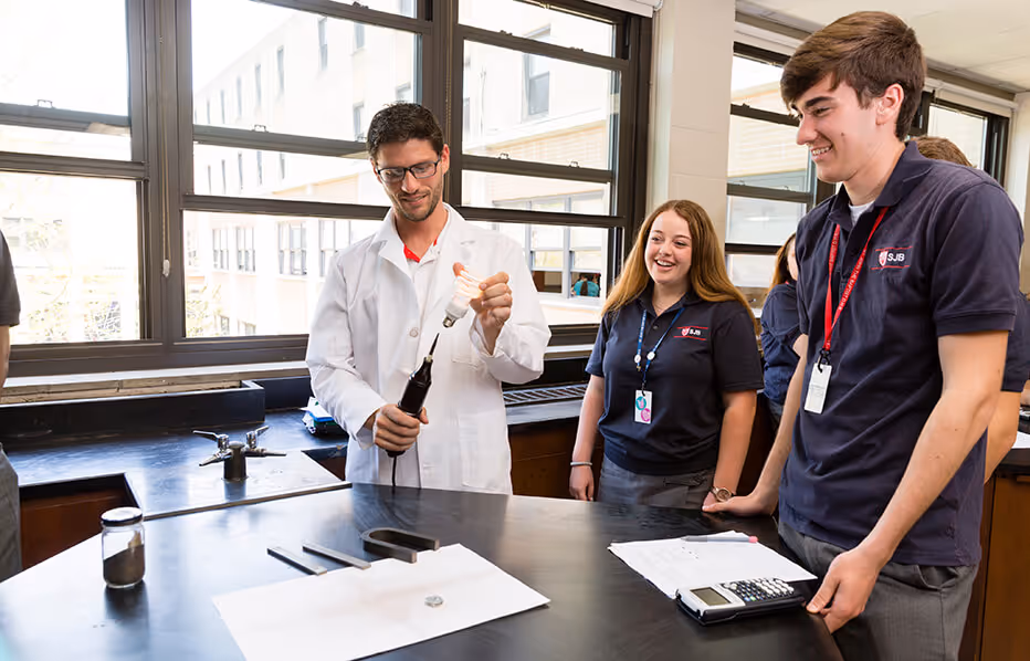 A teacher in a white lab coat demonstrating a light bulb to three smiling students in dark blue school uniforms in a bright classroom.