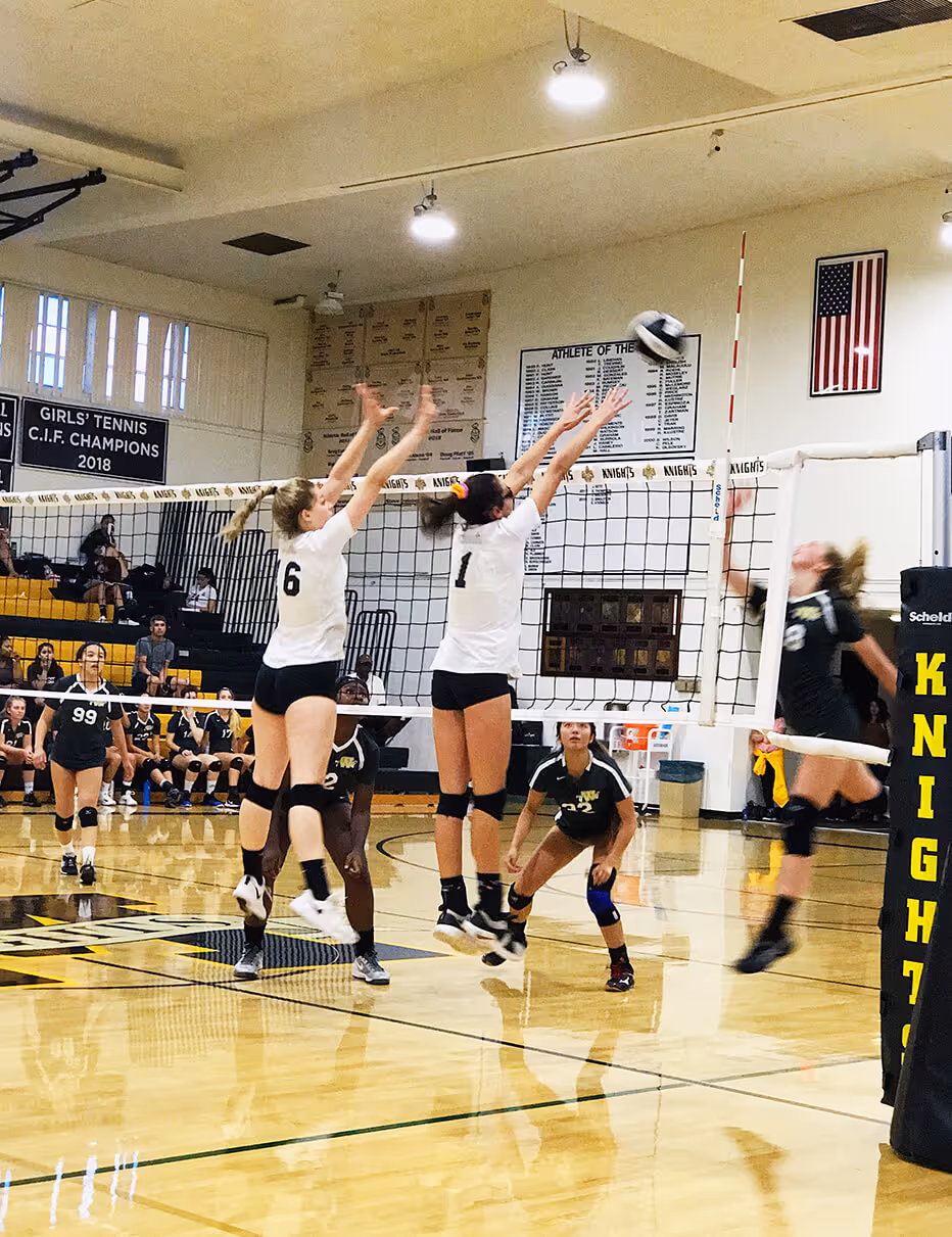 Two volleyball players in white jerseys jumping to block a ball at the net during a match with opposing players in black jerseys.