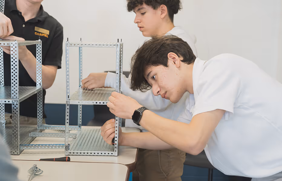 Three students assembling metal framework structures at a classroom table.