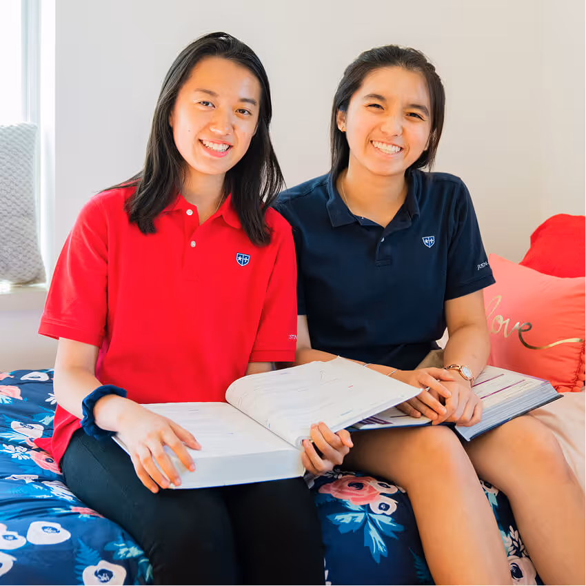 Two young women sitting on a bed with floral bedding, smiling and holding open notebooks for studying.