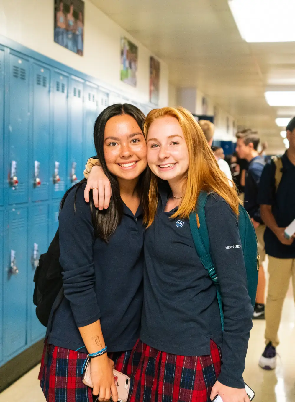 Two smiling schoolgirls in navy tops and plaid skirts hugging in a hallway with blue lockers.