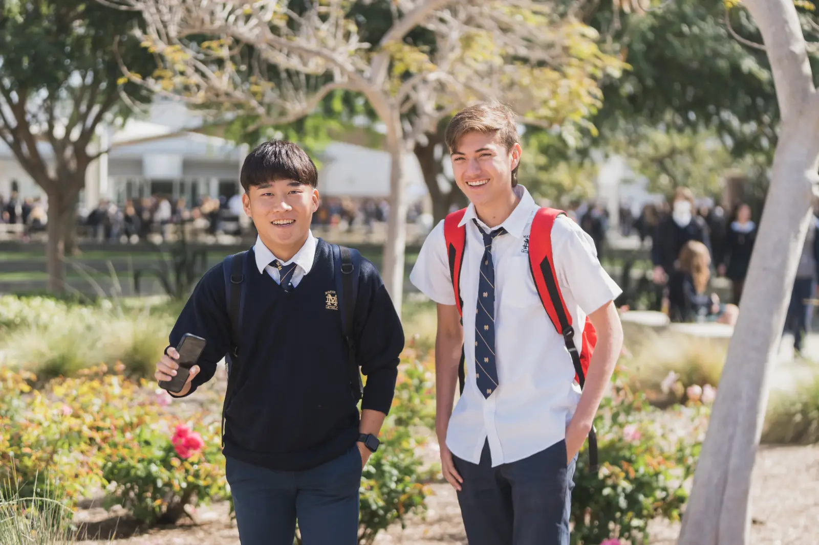 Two high school students in uniforms outdoors smiling, one holding a phone, backpacks on.