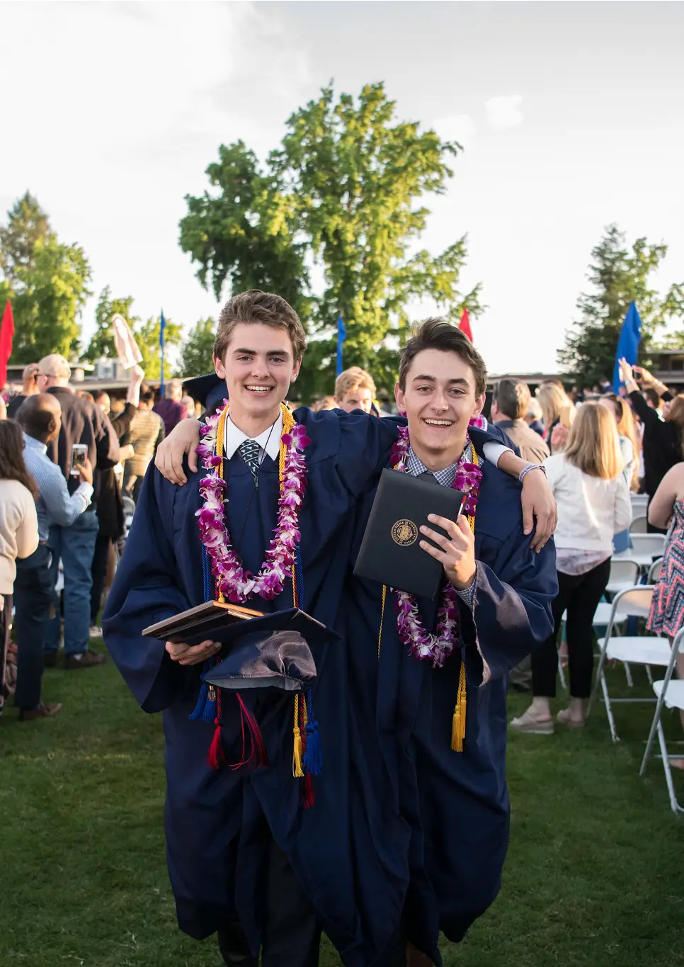 Two male graduates in blue gowns with flower leis smiling, holding diplomas at outdoor ceremony.