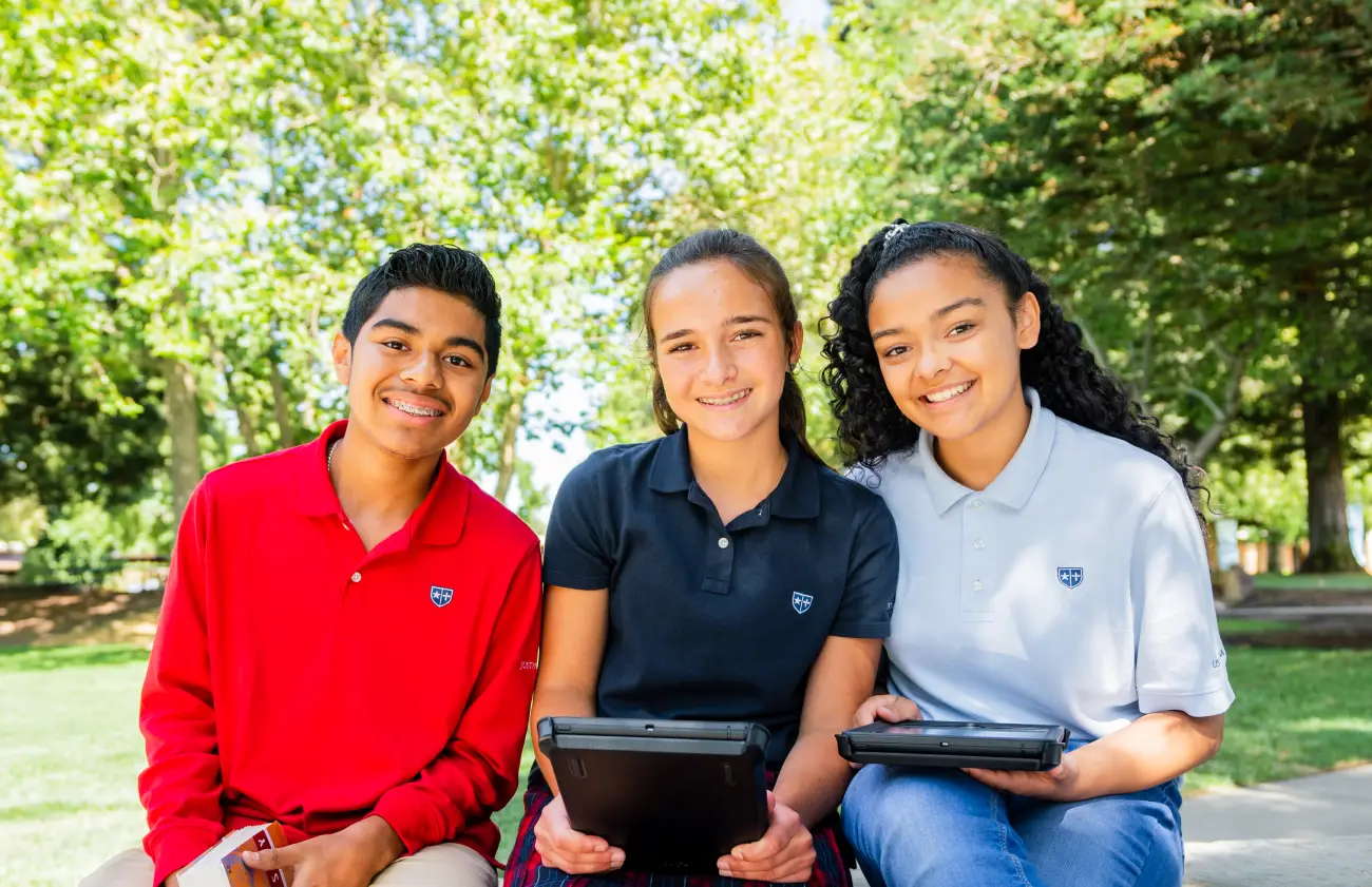 Three smiling teenagers in school uniforms sitting outside holding tablets with trees in the background.