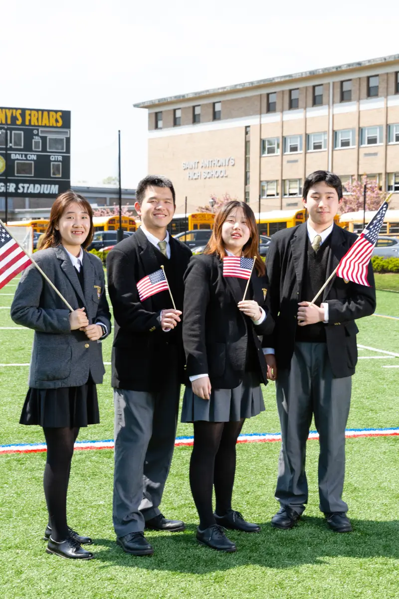 Four high school students in uniforms holding American flags on a football field at Saint Anthony's High School.