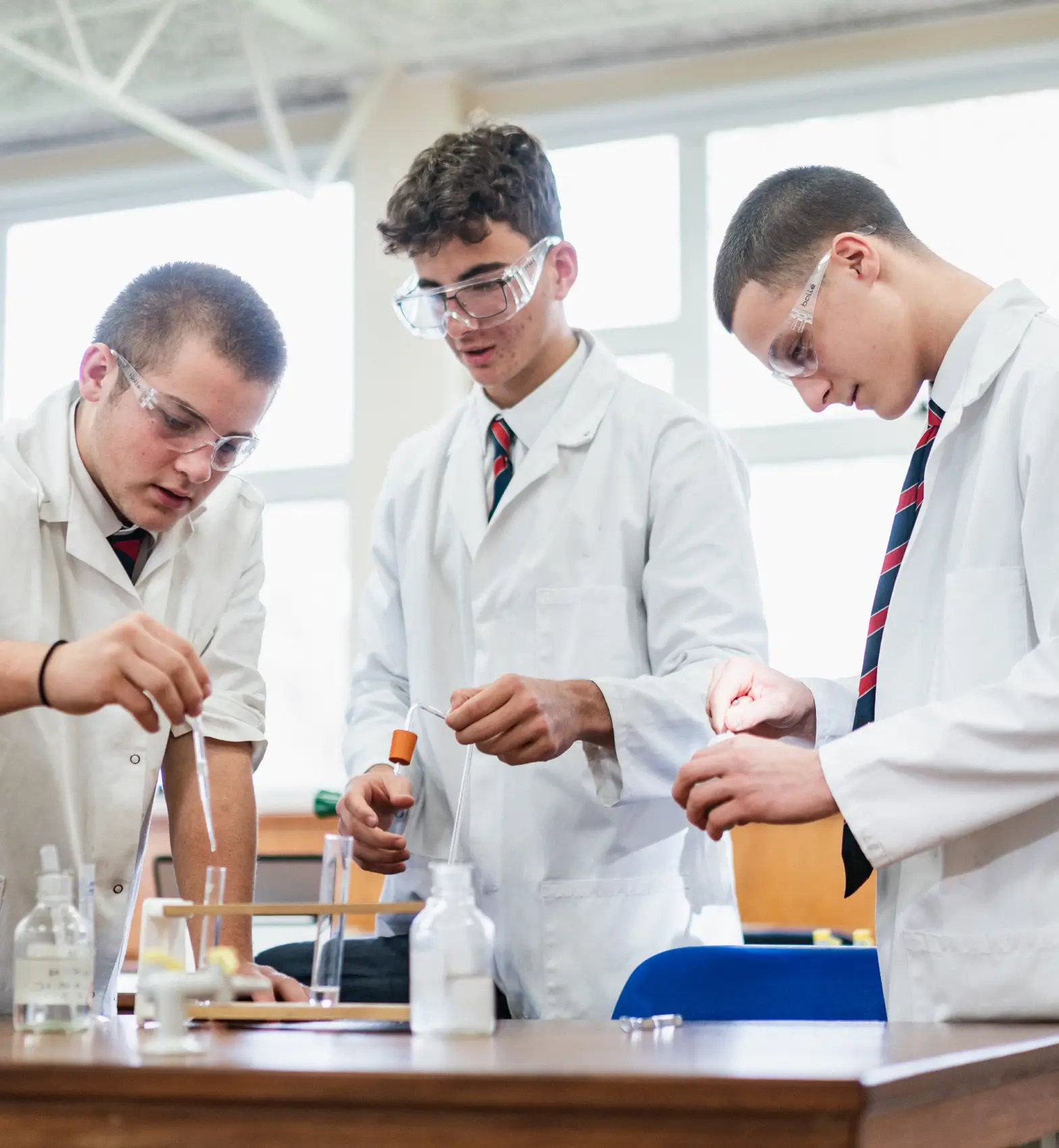 Three male students wearing lab coats and safety glasses conducting experiments with test tubes in a science lab.