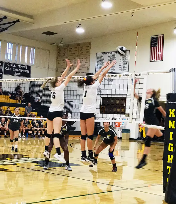 Two female volleyball players in white jerseys with numbers 6 and 1 jumping with arms raised to block a ball hit by an opposing player in black on the other side of the net in a gymnasium with banners and an American flag on the wall.