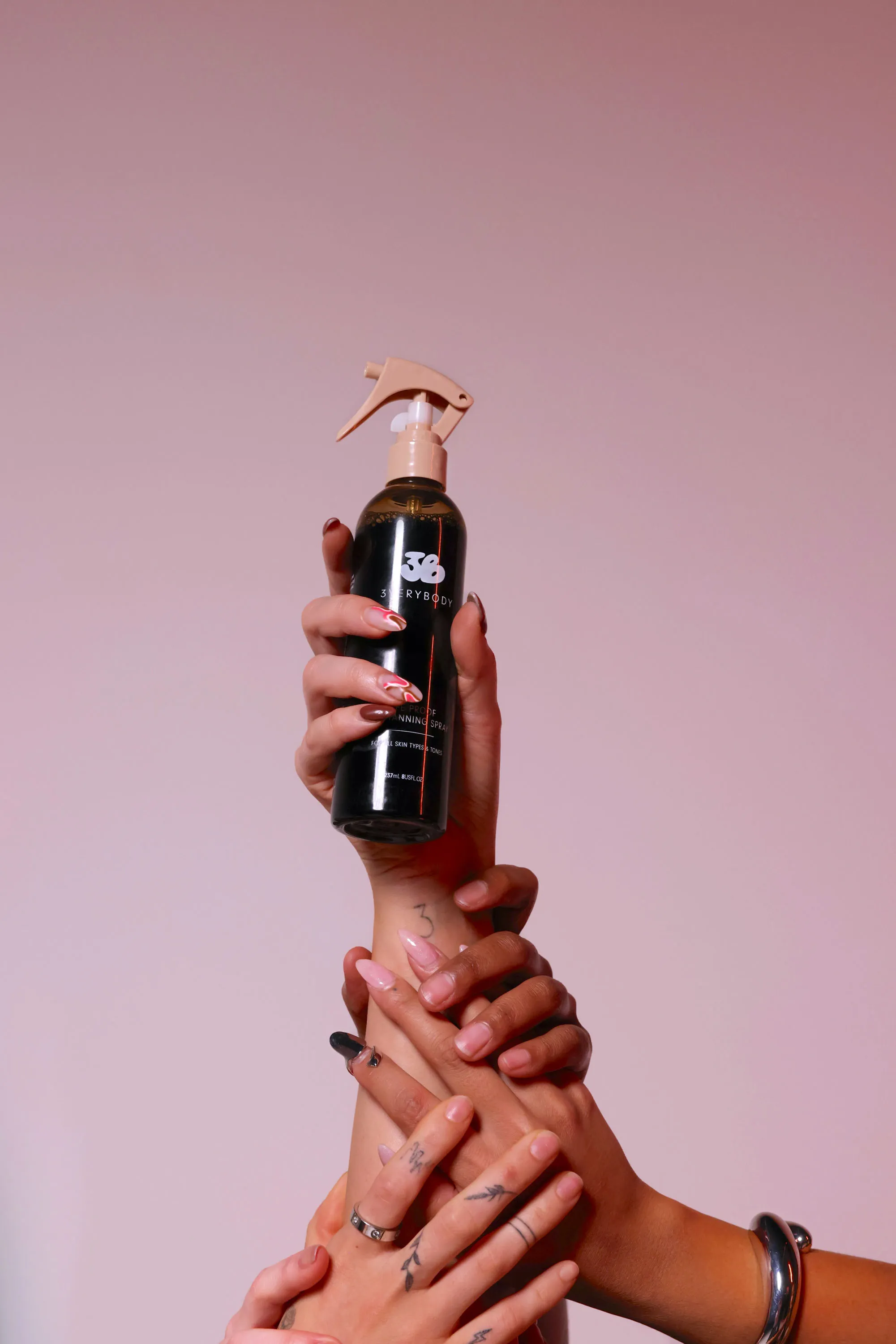 Multiple hands holding a black spray bottle with pink background and varied nail art.
