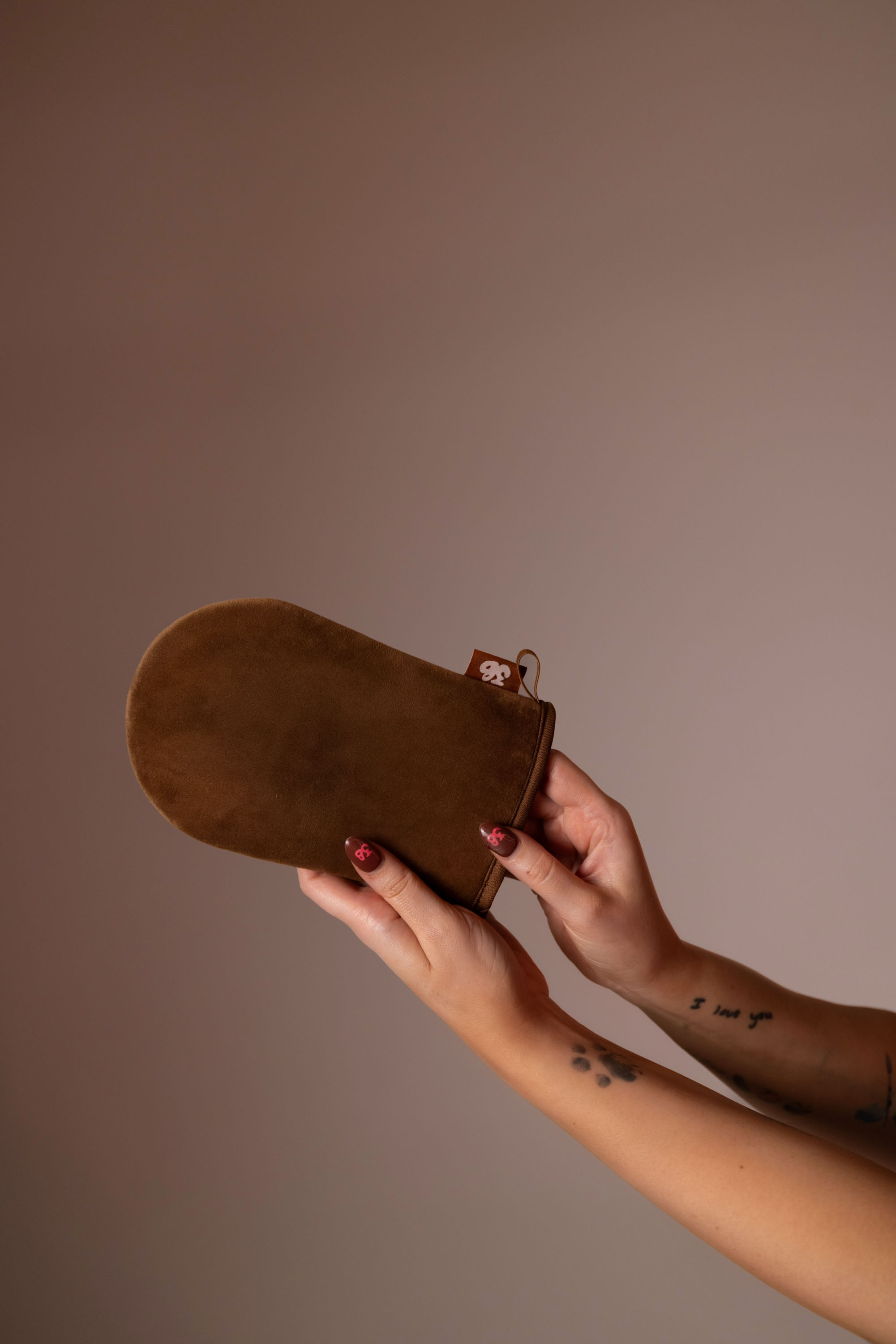 Two hands holding a brown 3VERYBODY tanning mitt