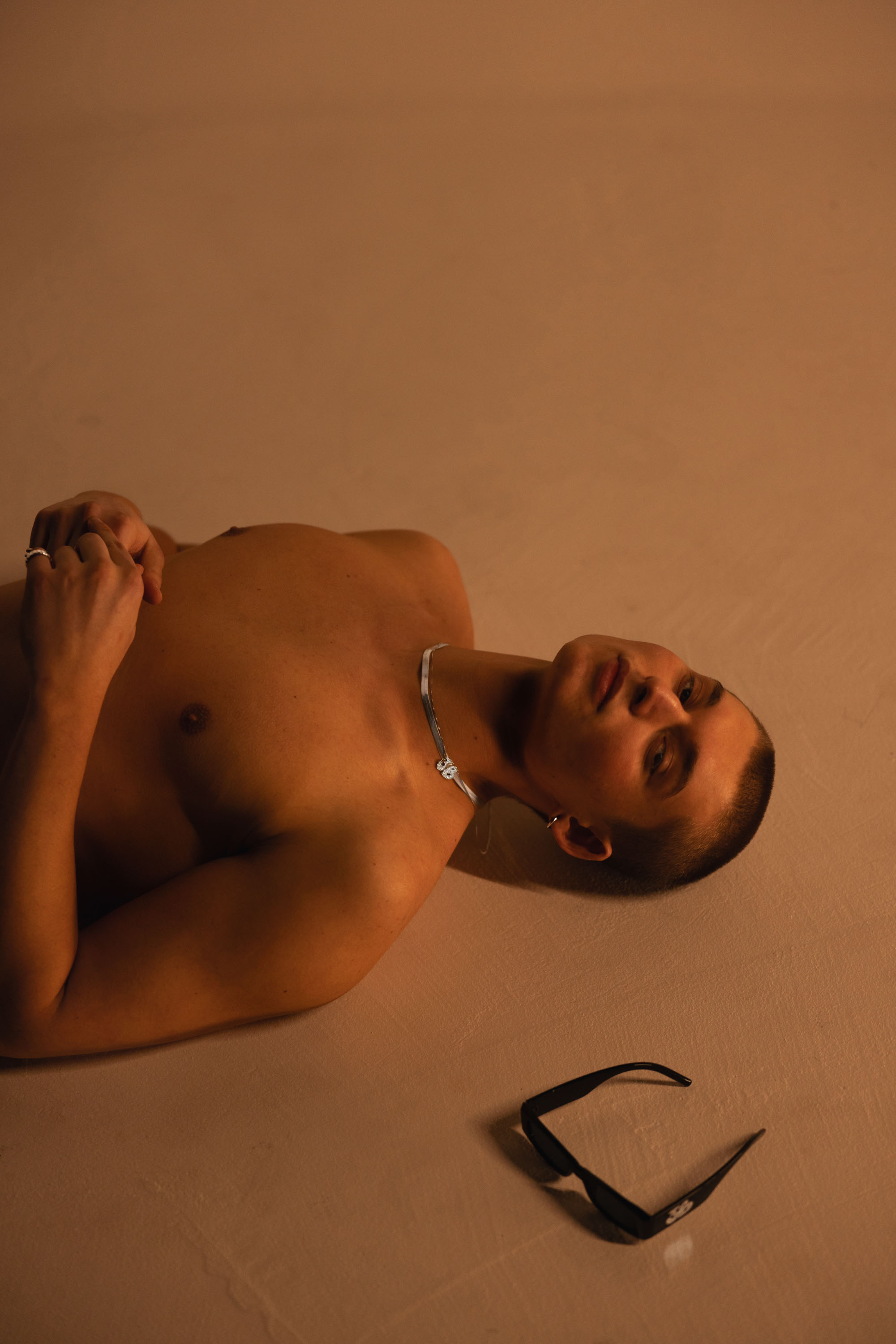 Man with a self tan lying on a tan backdrop next to a pair of sunglasses