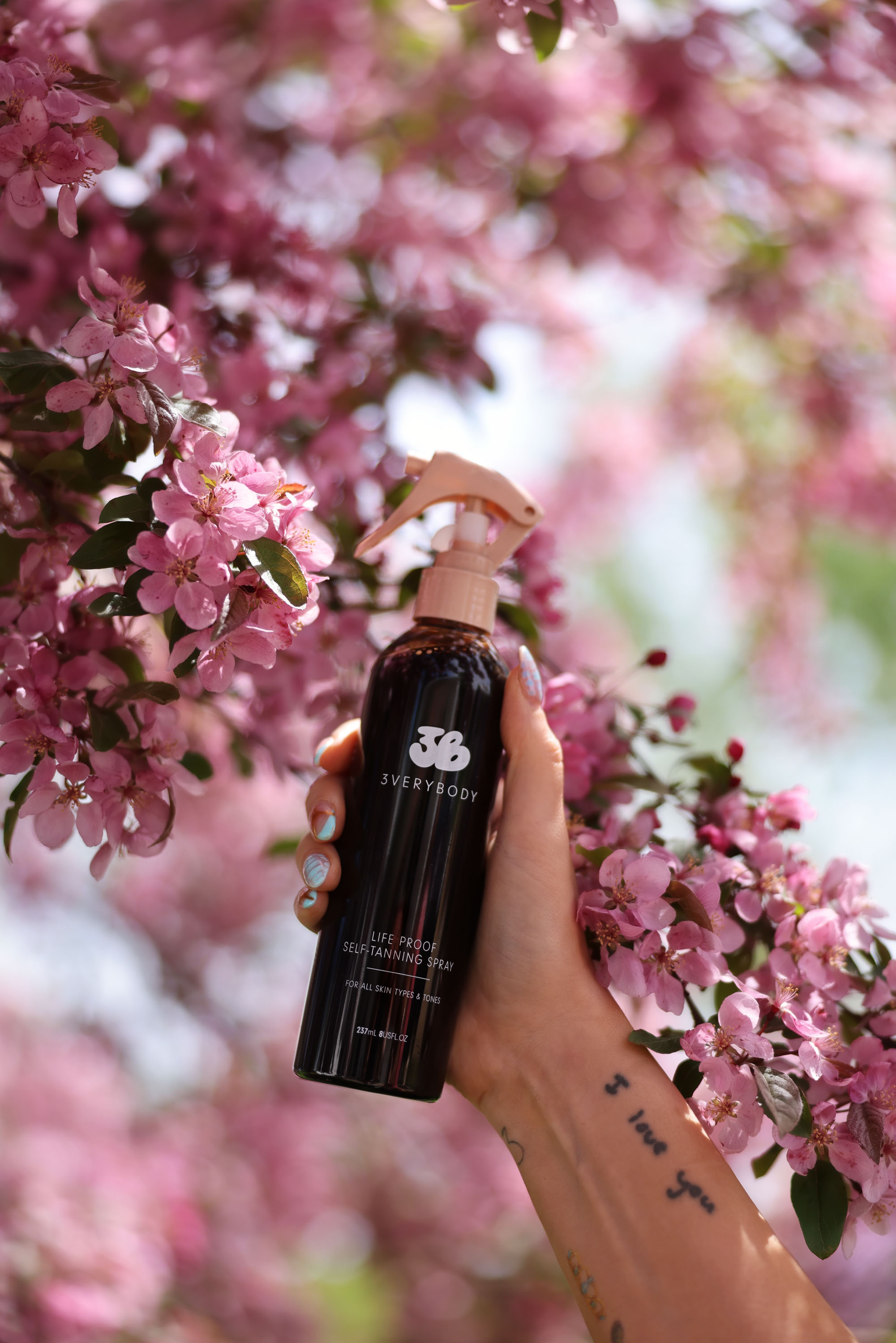 Hand holding 3VERYBODY Life Proof Tan spray among pink spring blossoms