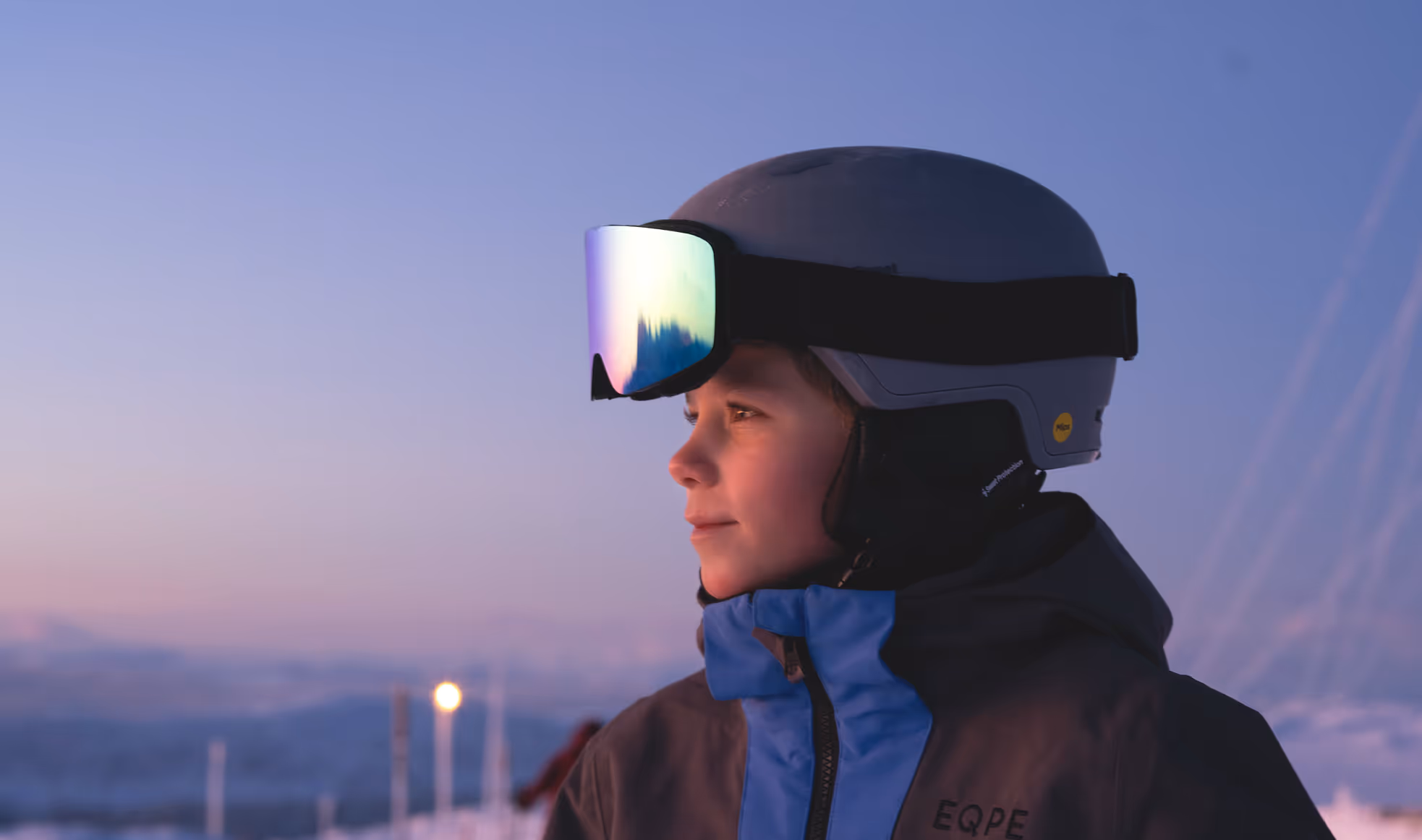 Närbild av en skidåkare med hjälm och stora skidglasögon, mot en skymningshimmel i mjuka, dova färgtoner