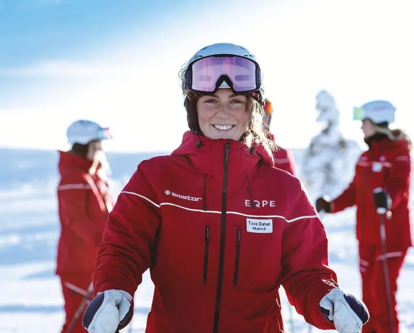 Leende SkiStar-skidlärare i röd jacka med hjälm och skidglasögon på snöigt fjäll med kollegor i bakgrunden