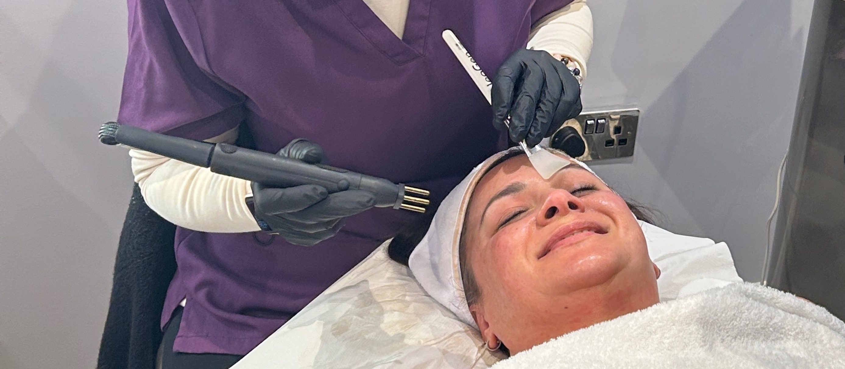 A woman receiving a facial treatment with a handheld device from a professional wearing gloves in a spa clinic.