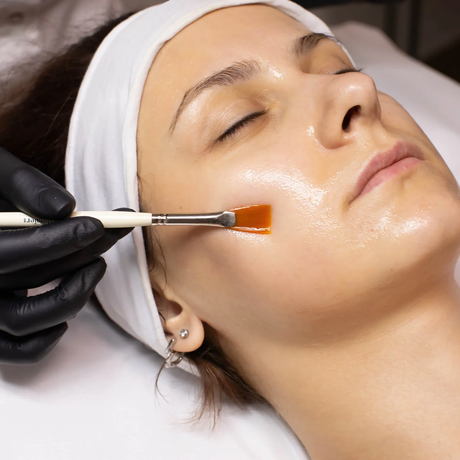 Close-up of a woman receiving a facial treatment with an orange peel applied to her cheek using a brush.