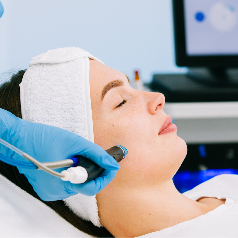 Woman receiving a facial treatment with a handheld device by a person wearing blue gloves and a white headband.