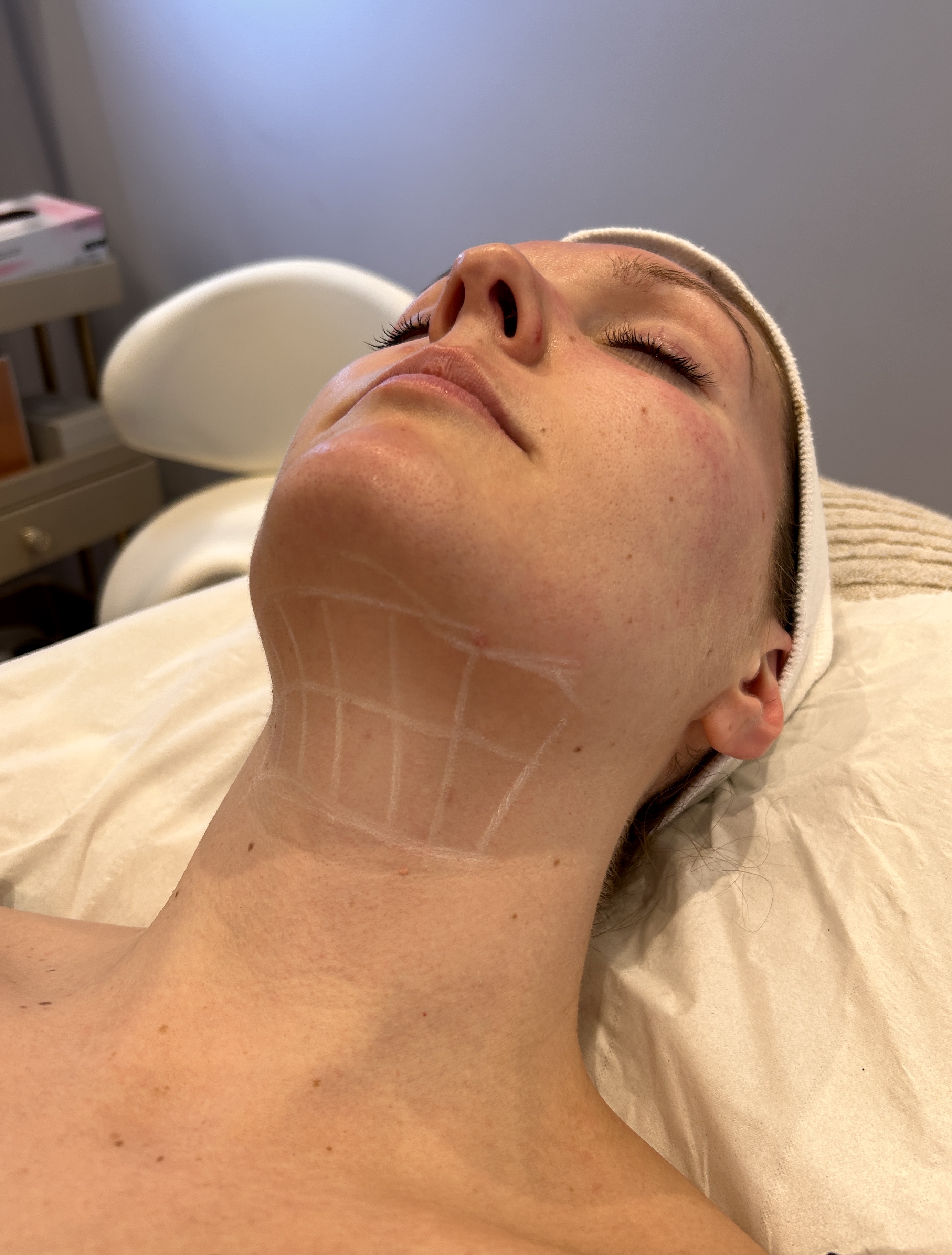 Woman lying down with under-eye gel patches receiving a facial treatment with a brush.