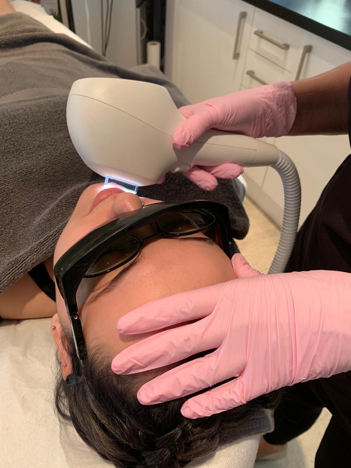 Woman lying down with eyes closed receiving a facial mask application while wearing under-eye gel patches and a headband.