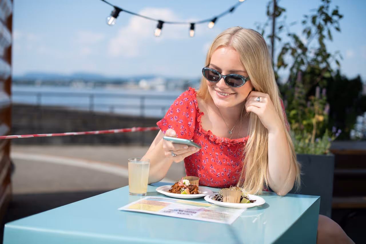 En smilende jente i 20 årene sitter på en uterestaurant og skanner en QR-kode på bordet