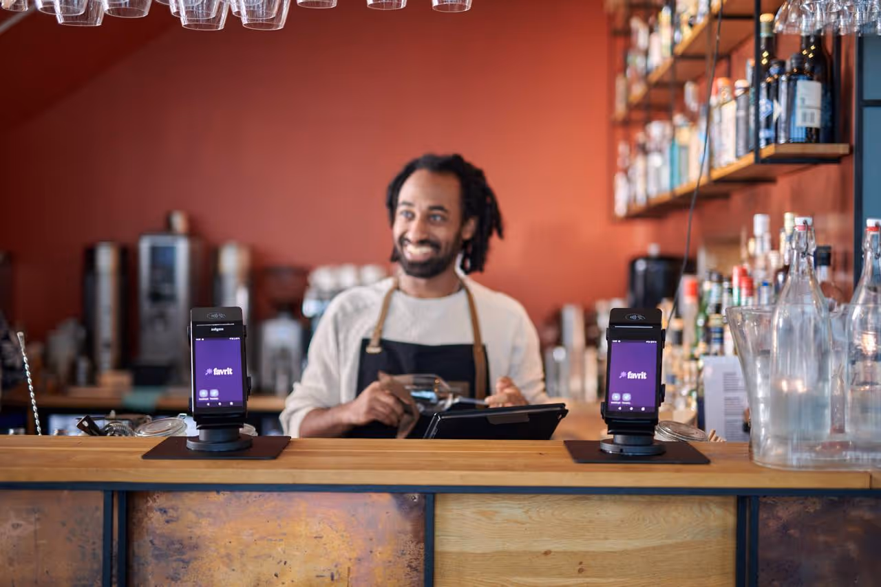 Smilende bartender med forkle bak en bar med to digitale betalingsterminaler med skjerm som viser appen Favrit.