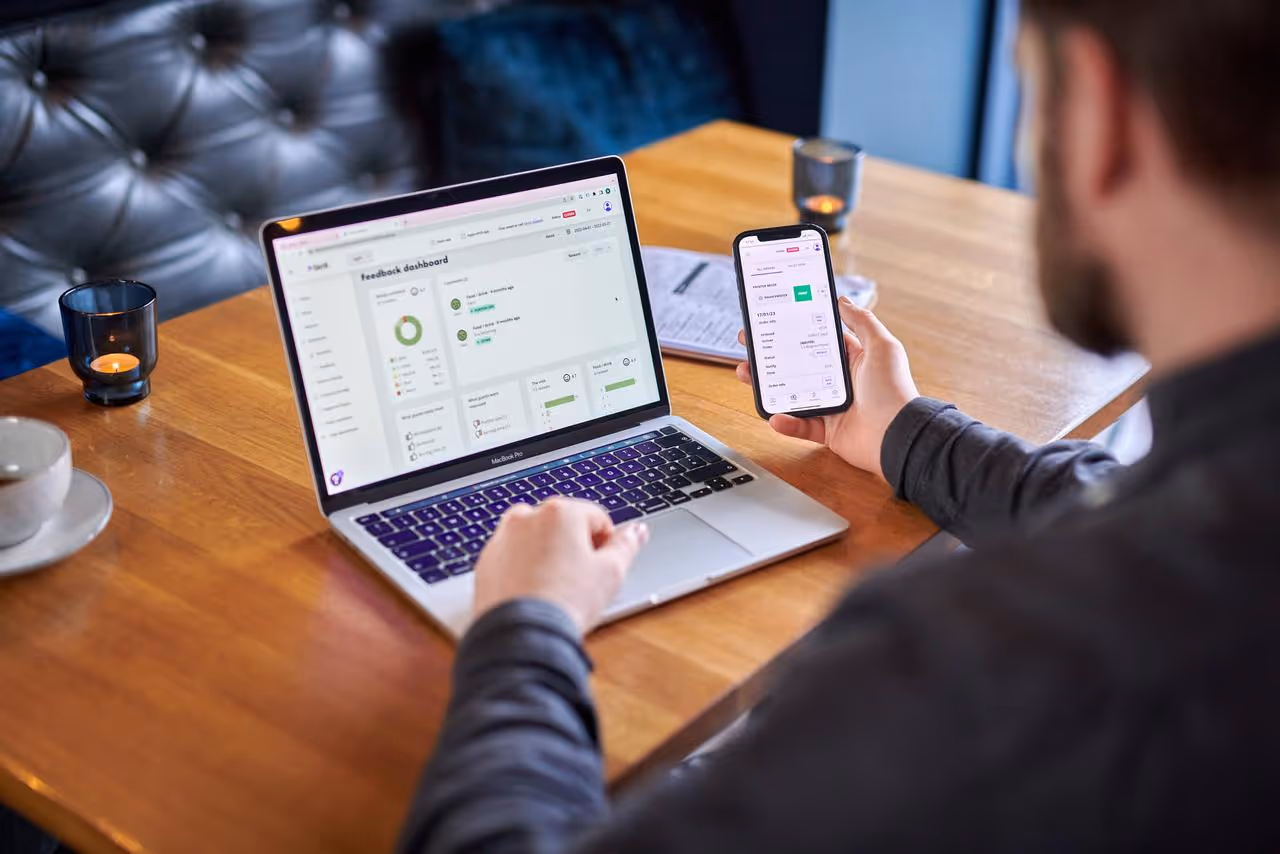 Person sitter ved et trebord med en MacBook Pro og følger med en app på mobilen med statistikk og tilbakemeldinger.