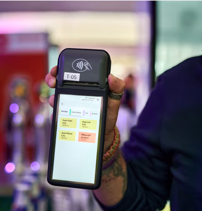 Hånd holder en mobil betalingsenhet med skjerm som viser ulike drikkekategorier og priser.