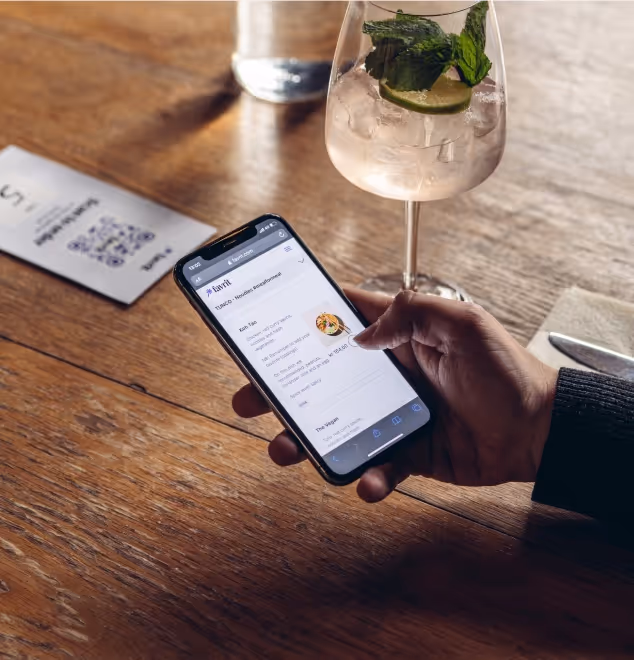 Hånd holder en mobiltelefon med en nettmeny for bestilling, plassert ved siden av et glass med iskald drikke på et trebord.