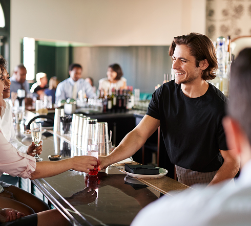 Smilende bartender strekker en drink til en kunde over en bar i en livlig restaurant.