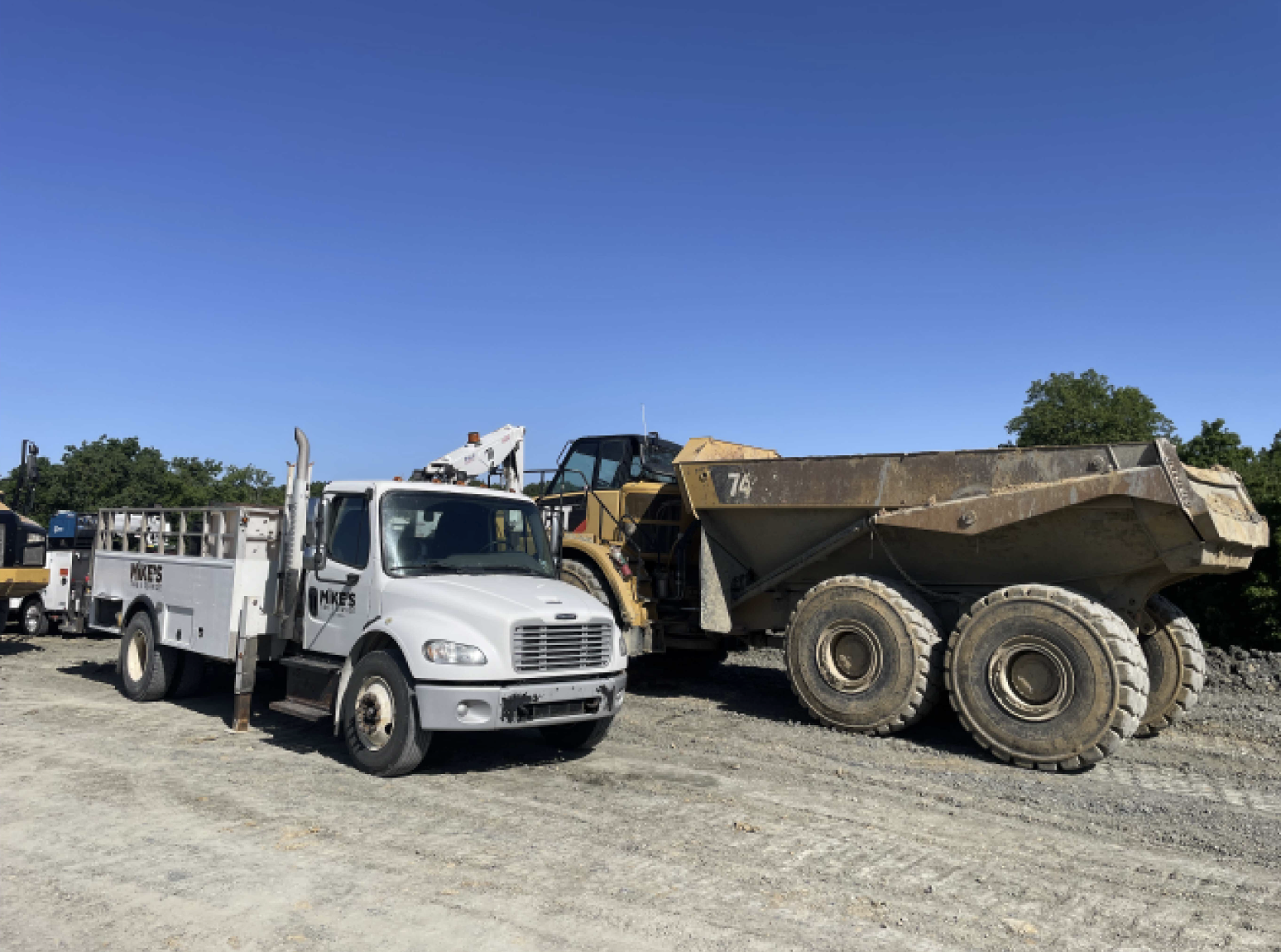 White utility truck with a crane attachment parked beside a large yellow dump truck on a gravel lot under a clear blue sky.