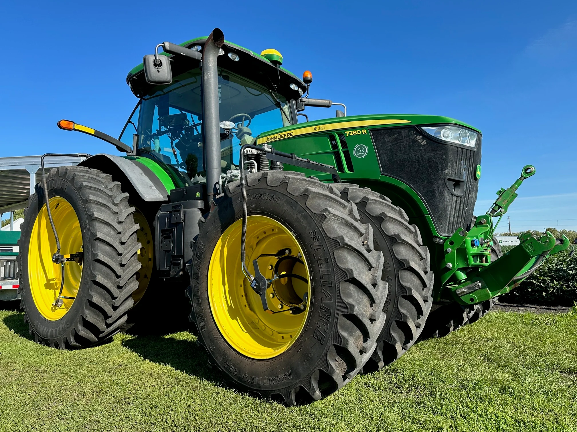 Green John Deere 7280R tractor with large yellow-rimmed dual front tires parked on grass under a clear blue sky.