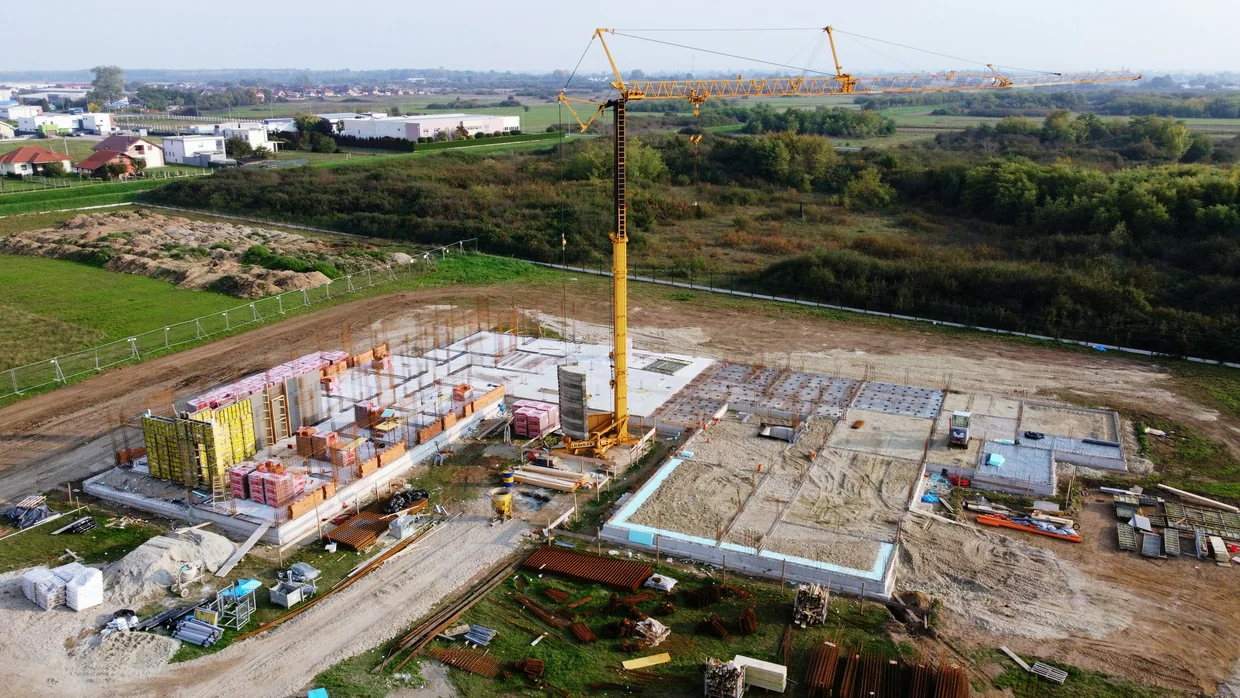 Aerial view of an early-stage construction site with a tower crane, concrete slabs and stacked materials surrounded by open fields