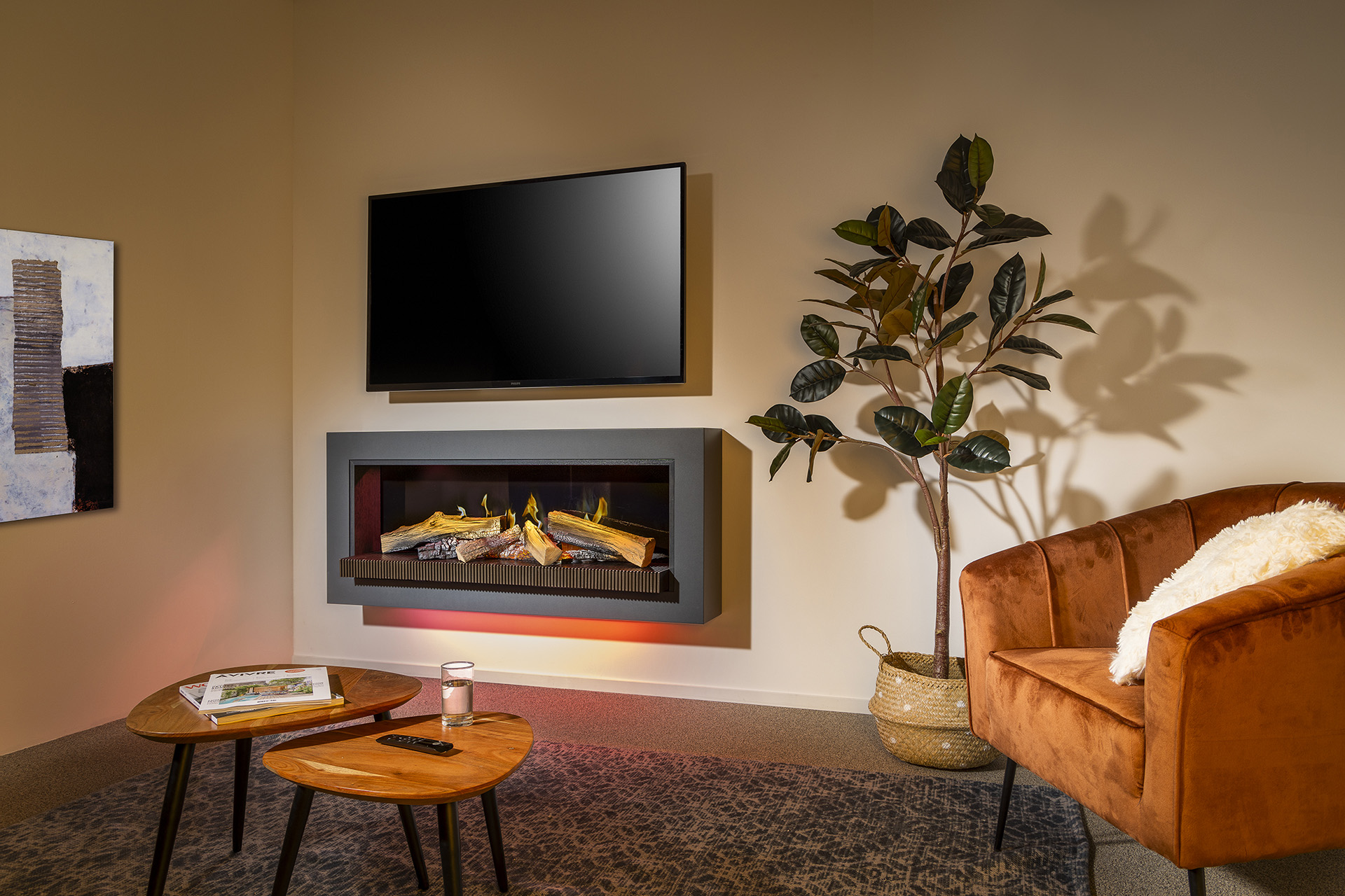 Image Alt Text Generator said:
Cozy modern living room interior with a wall-mounted flat-screen TV above a sleek electric fireplace featuring glowing faux logs and realistic flame effects. The space includes warm neutral walls, a rust-orange velvet accent chair with a white fluffy pillow, and a tall artificial rubber plant in a woven basket. In the foreground, two wooden nesting coffee tables sit on a patterned gray rug, one holding a glass of water, a magazine, and a TV remote. A contemporary abstract painting adds artistic flair to the left wall, while ambient lighting enhances the warm, inviting atmosphere—ideal for interior design inspiration, modern home decor, and electric fireplace setups.