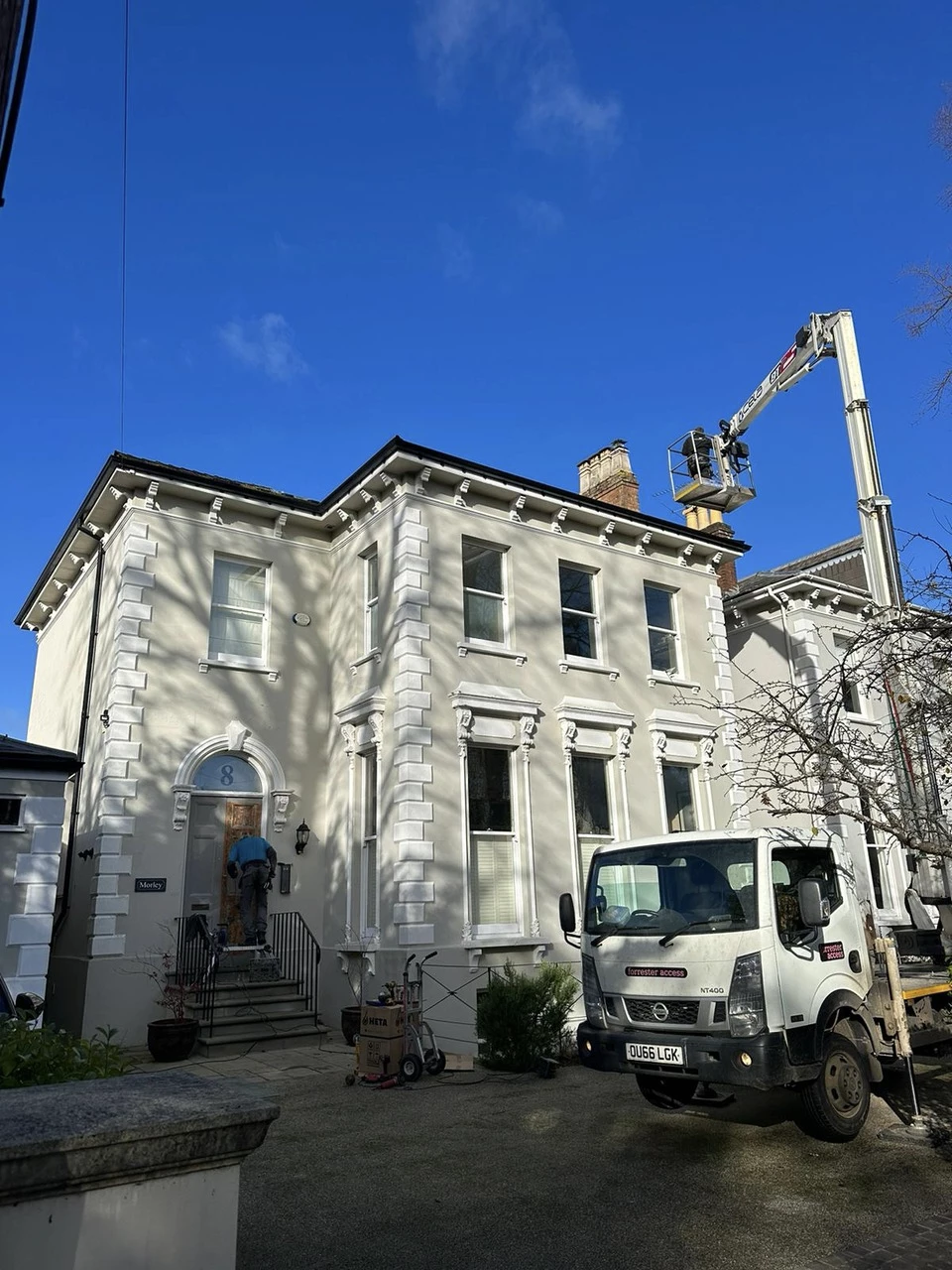 A white two-story house with a worker on a lift inspecting the chimney and a worker standing on the steps at the front door, with a white truck parked nearby.