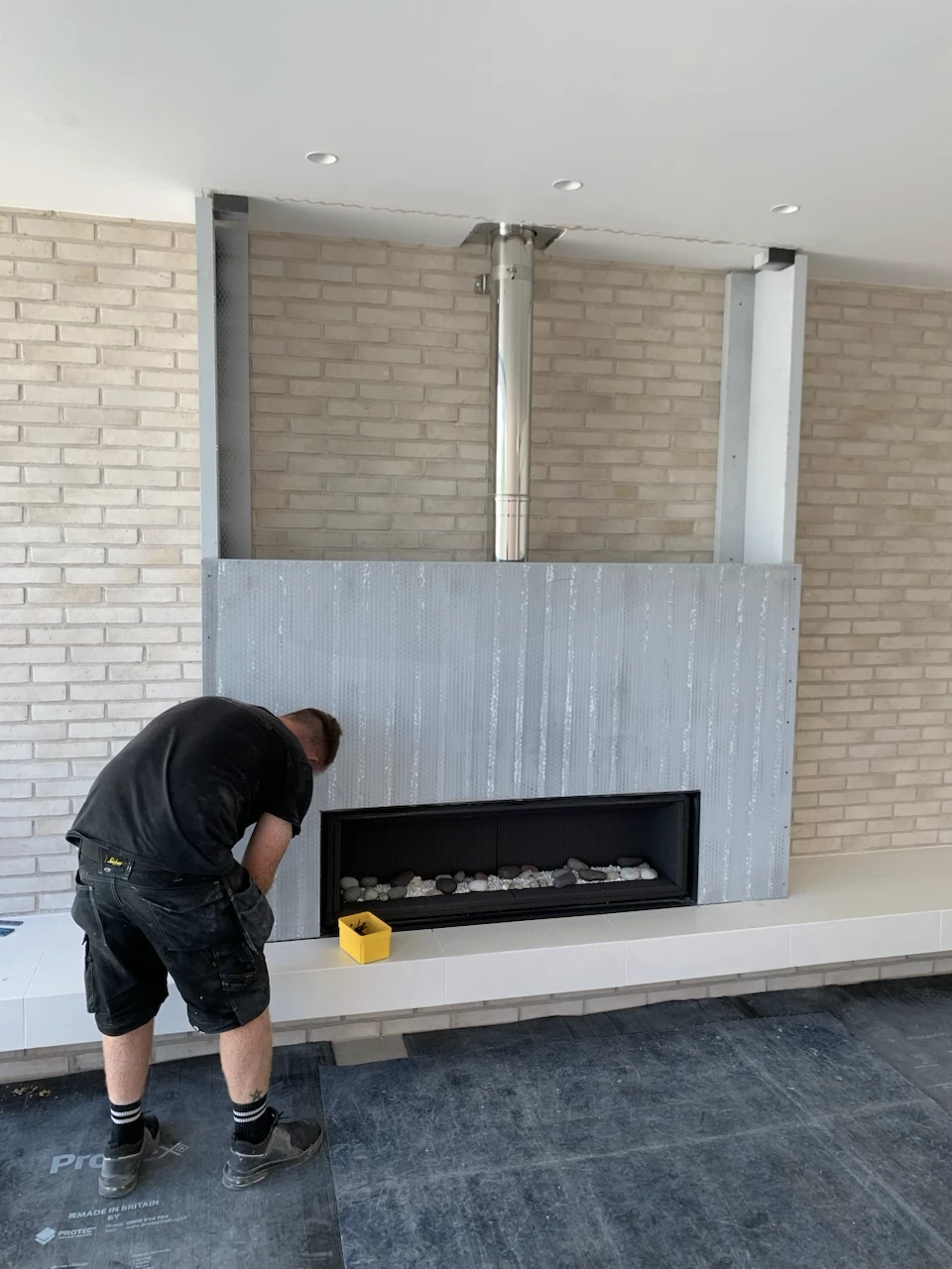 Worker installing or inspecting a modern inset fireplace with gray metal paneling and chimney pipe against a light brick wall.
