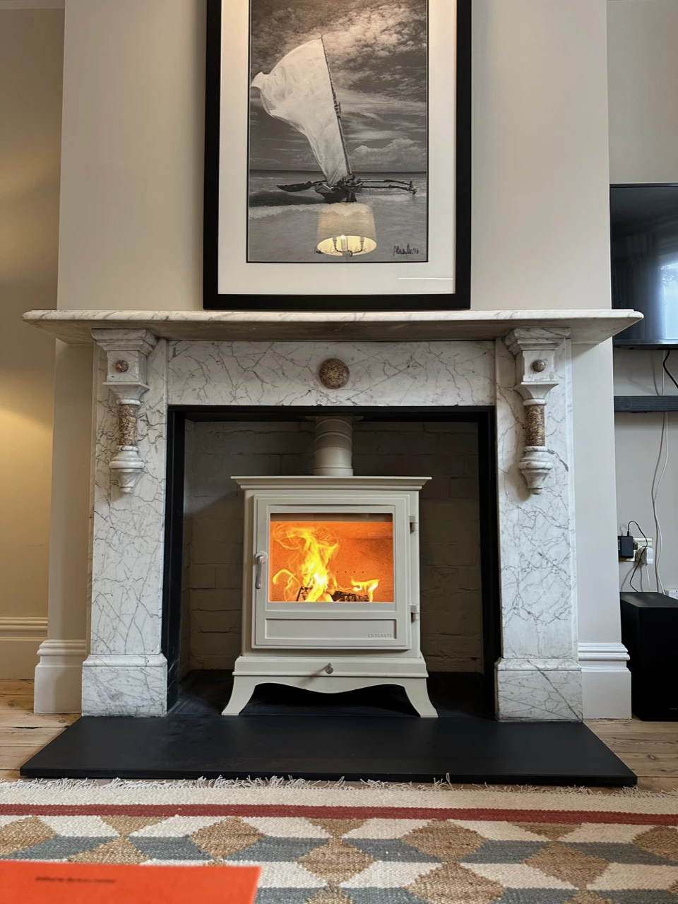 White wood-burning stove with visible flames inside, set within a marble fireplace, with a black hearth and a framed black-and-white sailboat photo above.