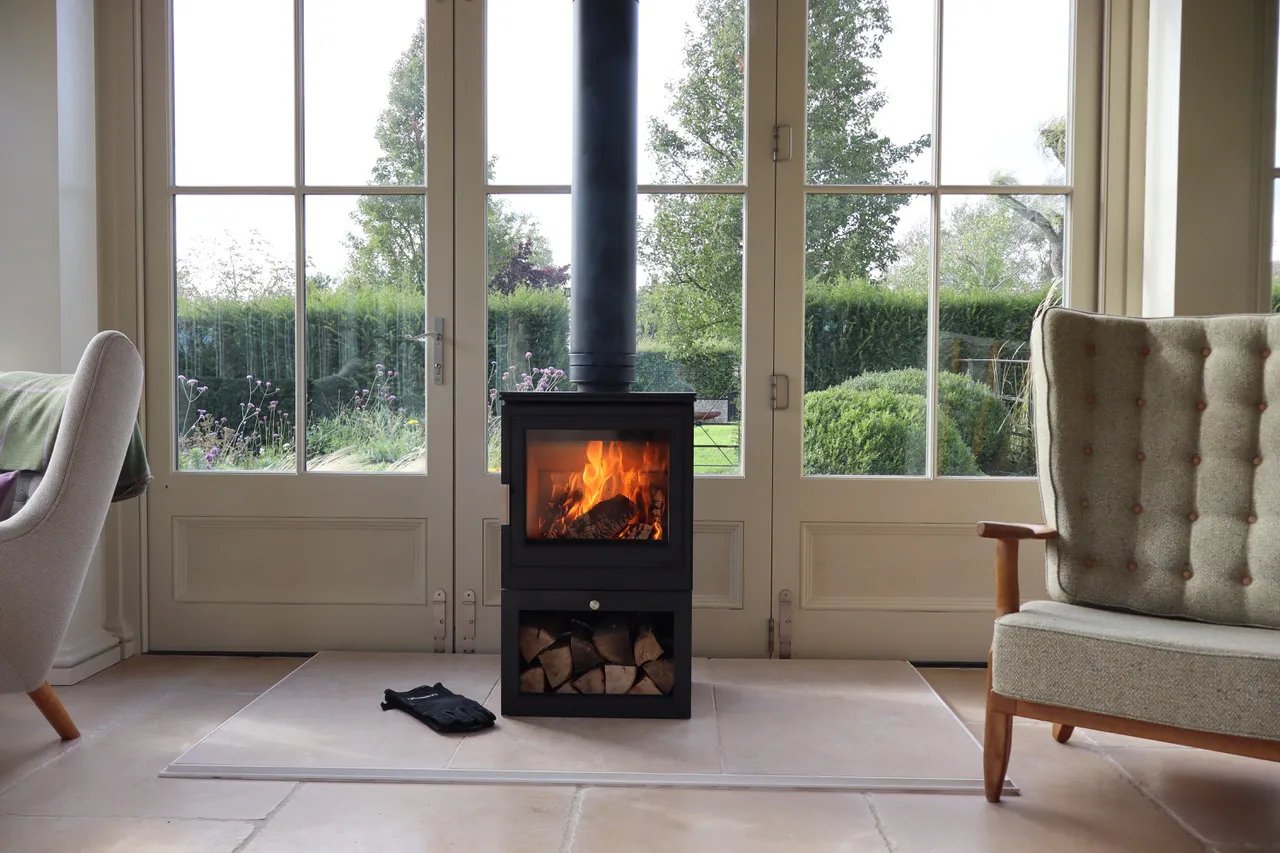 Modern black wood-burning stove with fire inside placed in front of large glass doors overlooking a green garden, with a glove on the floor and armchairs on each side.