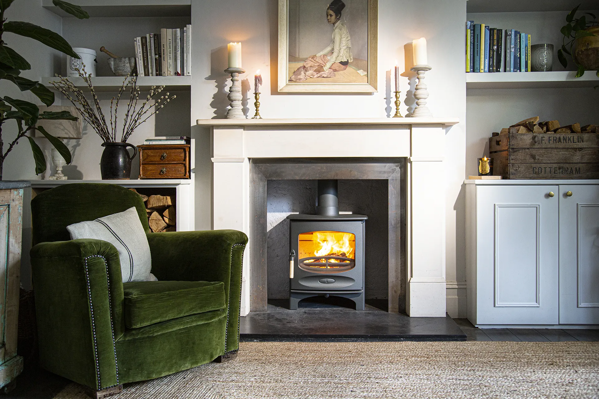 Cozy living room with green armchair, fireplace with burning stove, candles, books, and wooden crates.