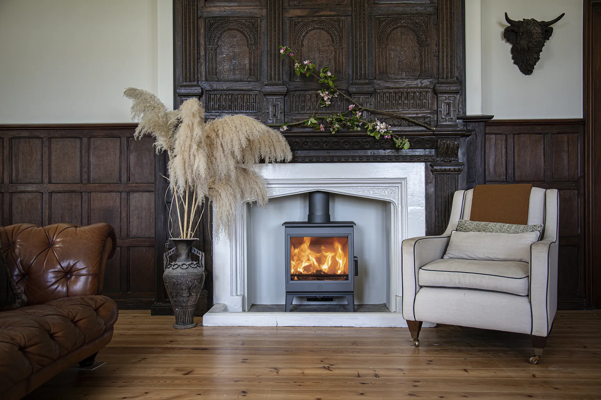 Living room with lit wood stove in white fireplace, brown leather sofa, beige armchair, and decorative vase.