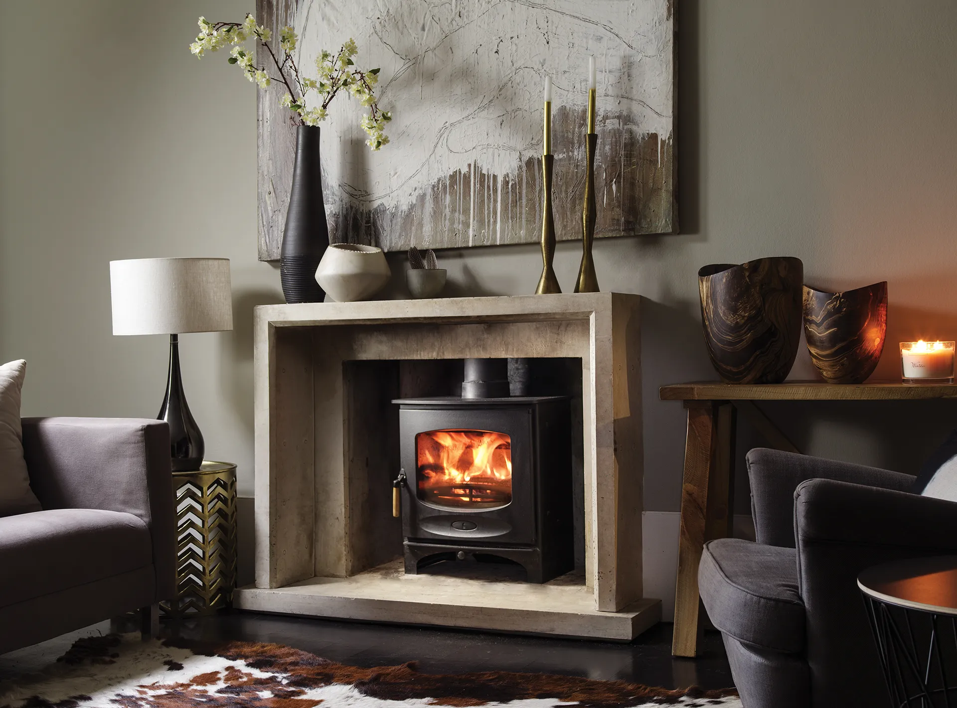 Cozy living room with burning fireplace, gray armchairs, a lamp, decorative vases, and wall art.