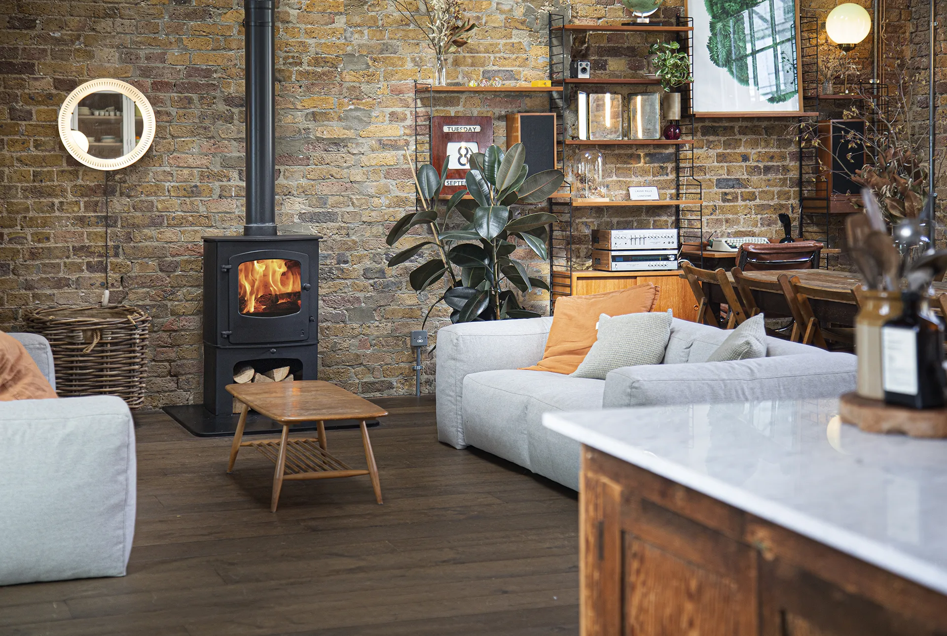 Cozy living room with a burning wood stove, gray sofas, wooden coffee table, plants, and exposed brick walls.