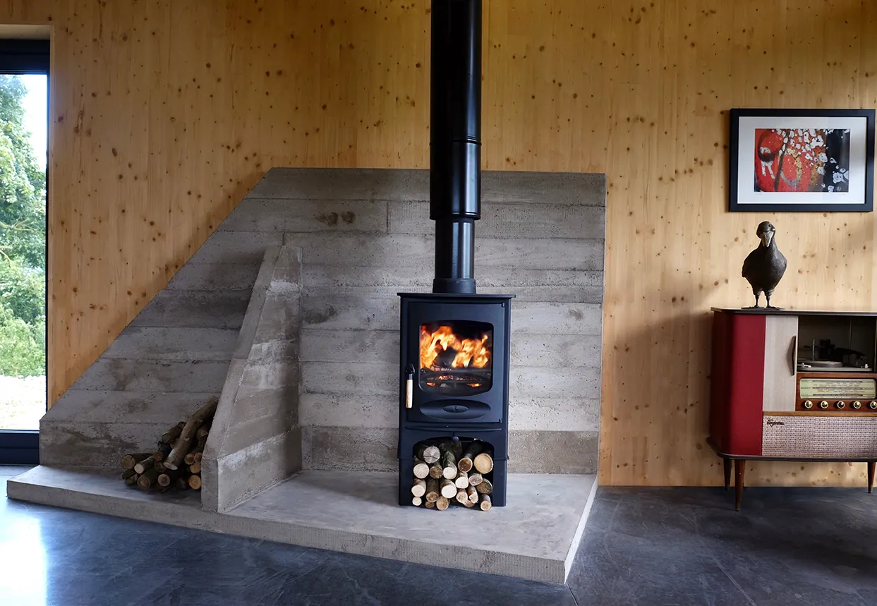 Black wood stove with fire burning and logs below in a modern room with wooden walls and concrete hearth.
