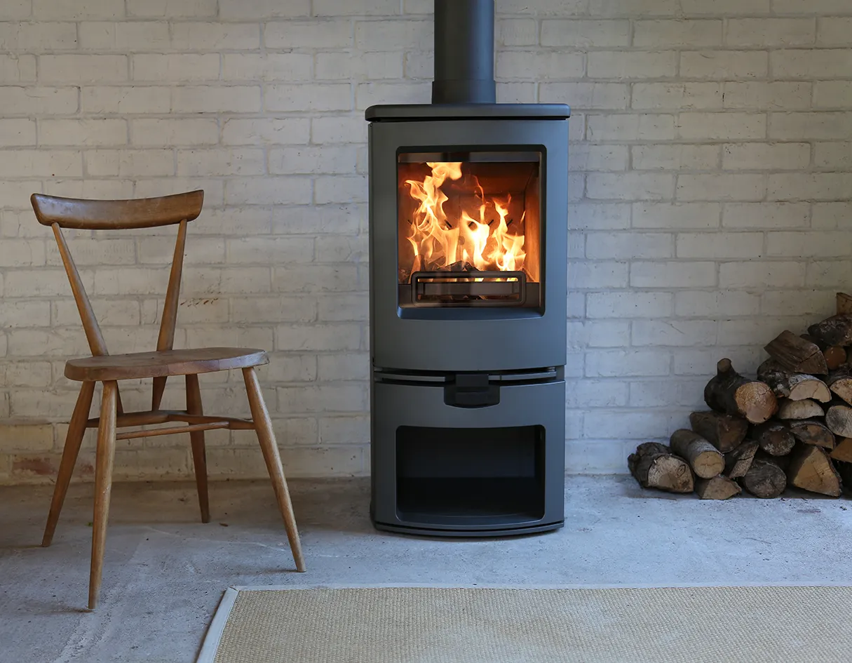 Modern gray wood-burning stove with fire inside next to a wooden chair and stack of logs.