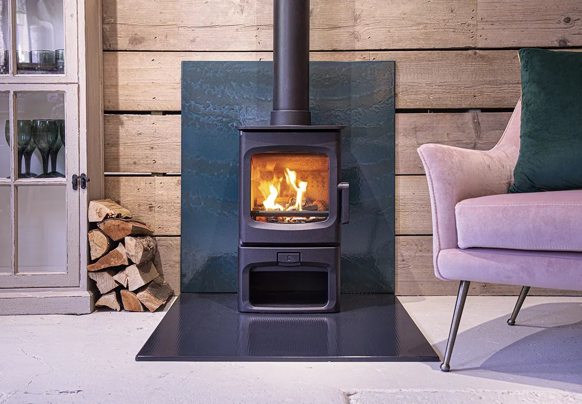 Modern black wood stove with fire burning, next to stacked firewood and a pink armchair with a green pillow.