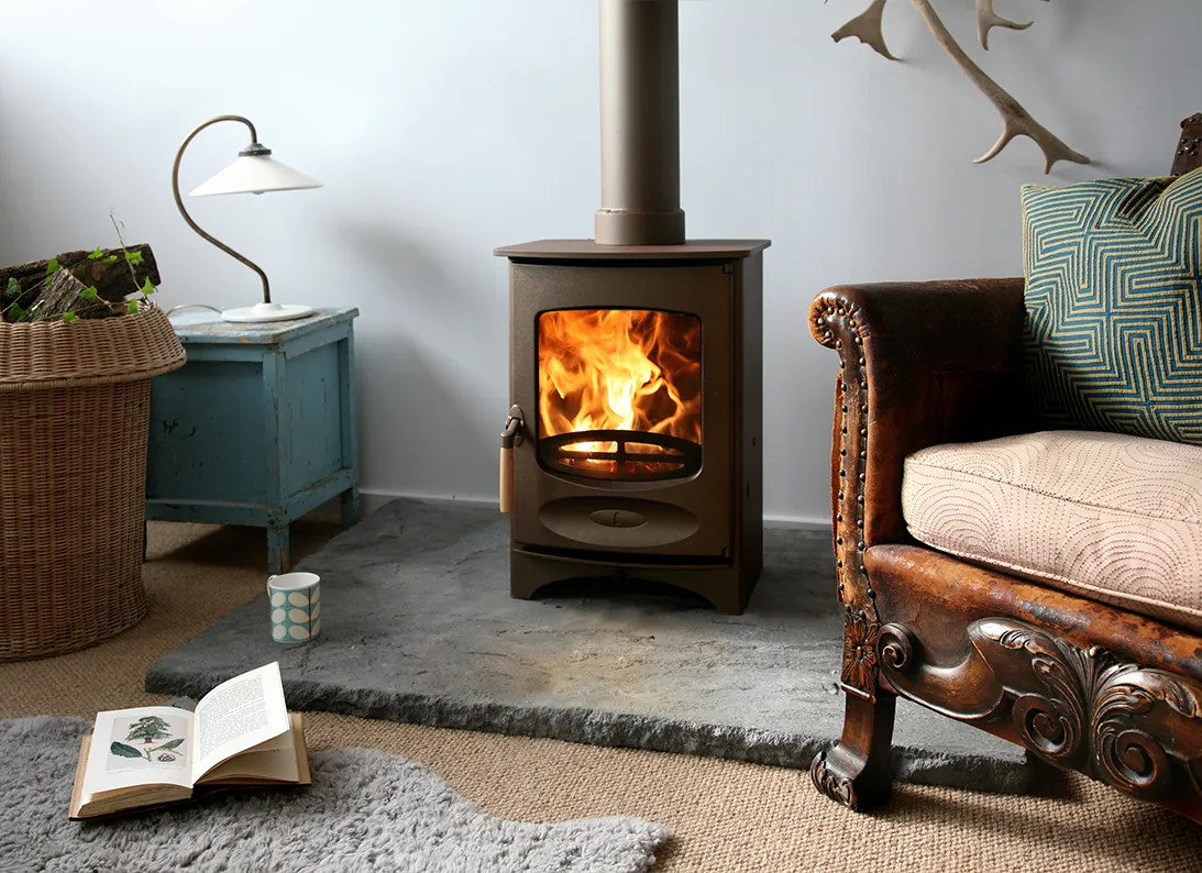 Burning wood stove on stone hearth between vintage chair and blue side table with lamp and basket.