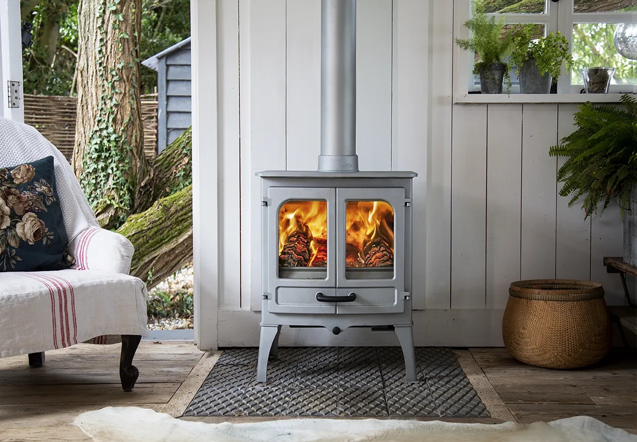 Pewter wood-burning stove with fire inside in a cozy room with white walls and wooden floor.
