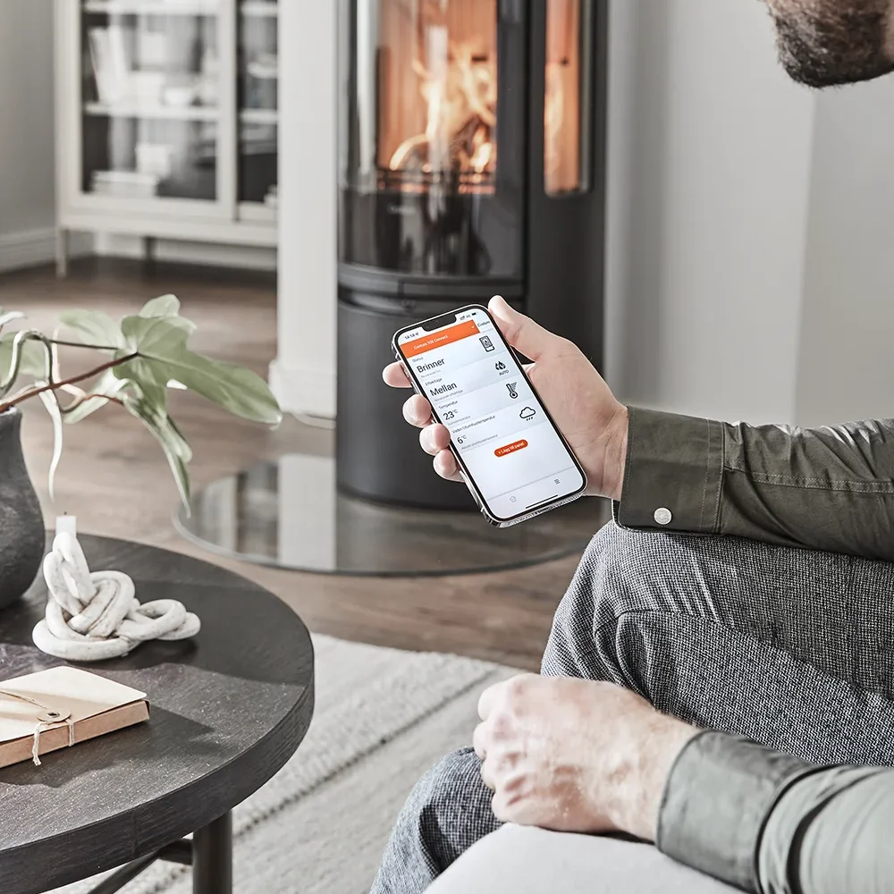 Person holding smartphone showing a fireplace control app with a burning stove in the background.