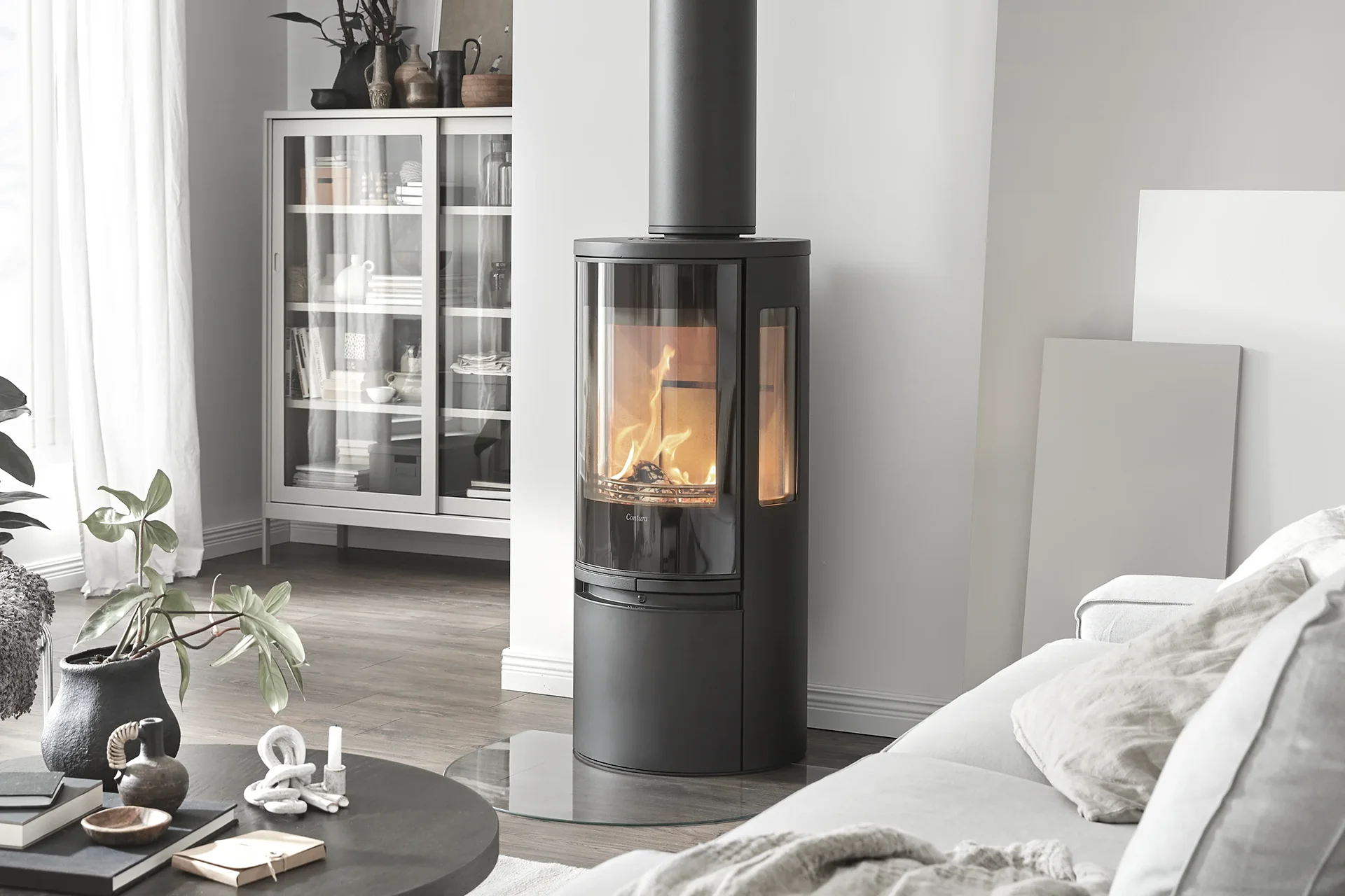 Modern black cylindrical wood stove with fire burning, set in a cozy living room with neutral decor.