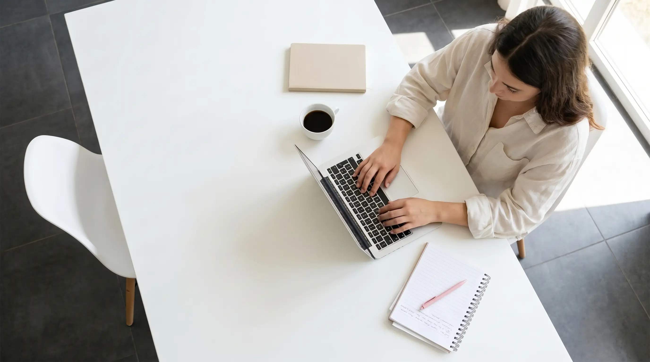 Femme tapant sur un ordinateur portable à une table blanche avec café et carnet de notes.