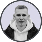 Portrait of a young man wearing a white hoodie and black jacket smiling against a light gray background.