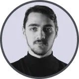 Portrait of a young woman with long dark hair and bangs wearing a black top, smiling slightly against a light purple background.